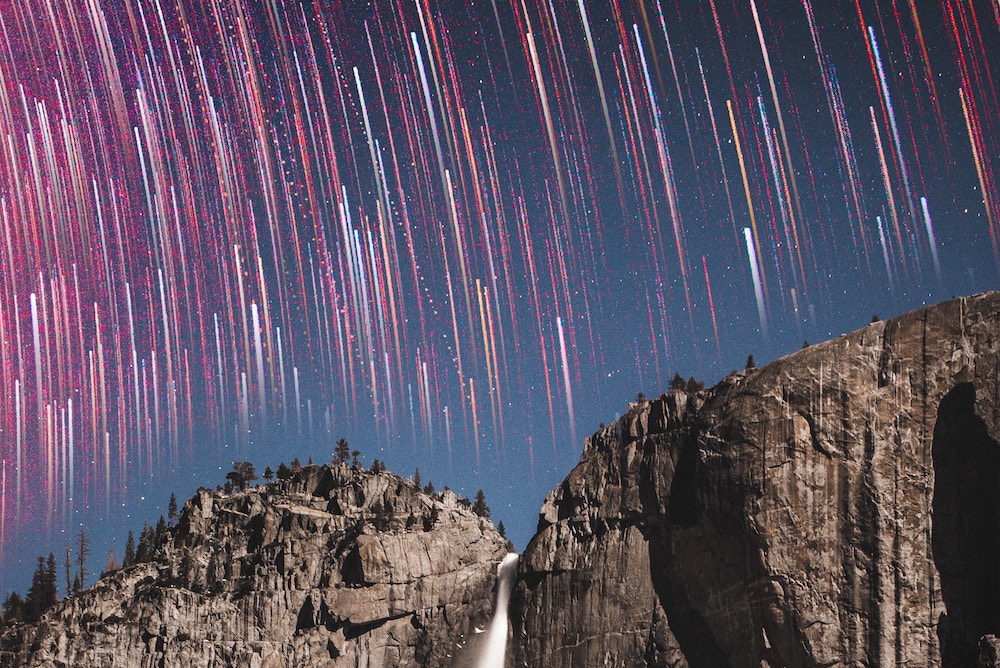 Long exposure of stars in the night sky in front of a waterfall and rock face.