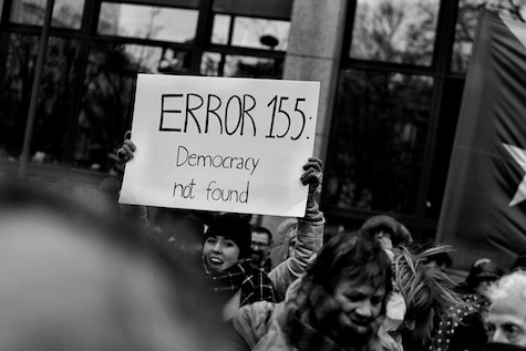 Person at a rally is holding a sign that reads "Error 155: Democracy Not Found". Photo by Randy Colas on Unsplash